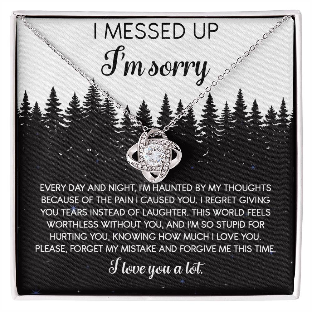 The Sorry-By My Thoughts - Love Knot Necklace, featuring a heart-shaped pendant adorned with cubic zirconia crystals, is displayed on a card with an apology message. The card reads: "I messed up. I'm sorry. Every day and night, I'm haunted...I love you a lot," set against a serene forest background.