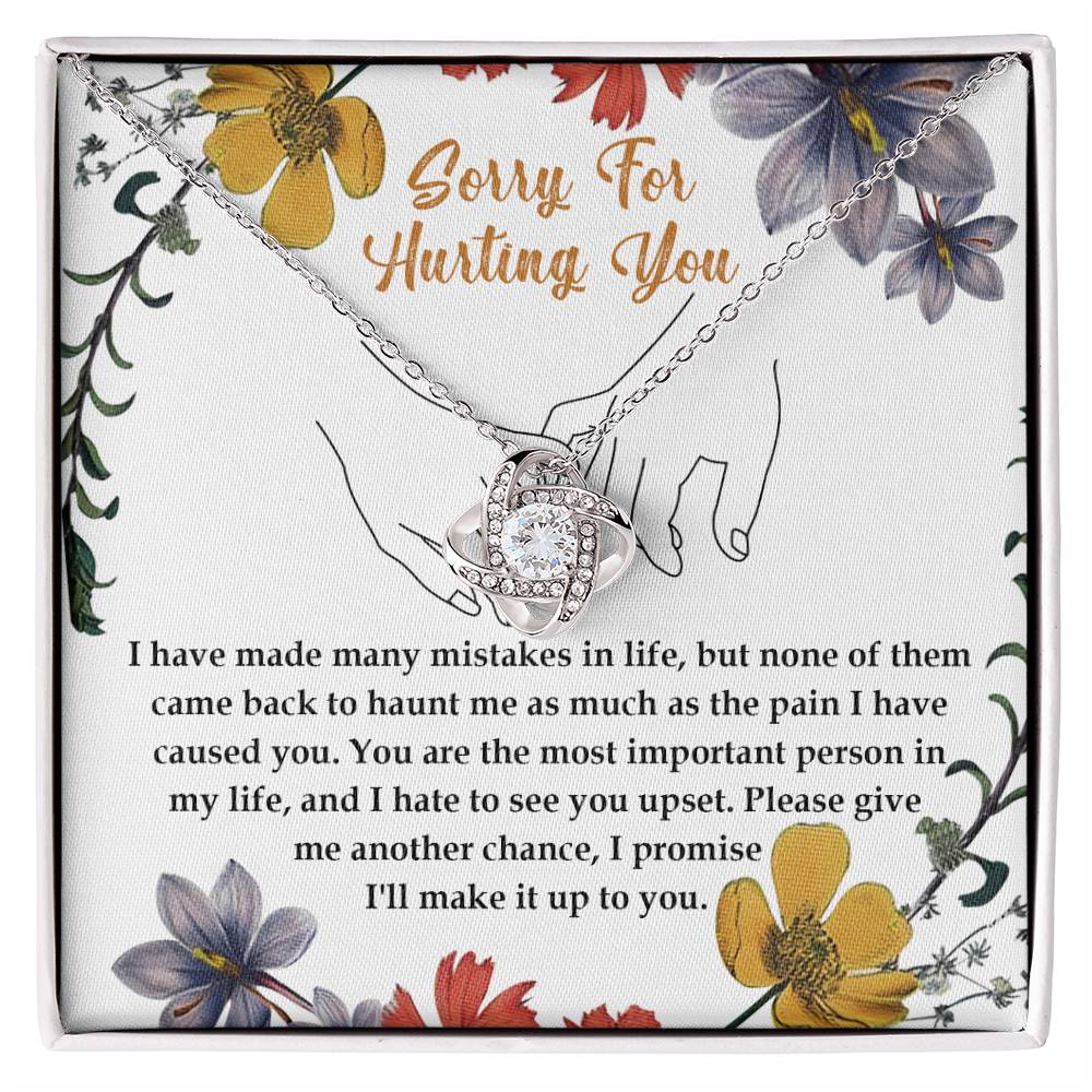 The Sorry-Haunt Me - Love Knot Necklace is a silver piece featuring an intertwined heart and infinity symbol adorned with cubic zirconia crystals, elegantly presented against a floral card that reads: "Sorry For Hurting You" with an apology message below.