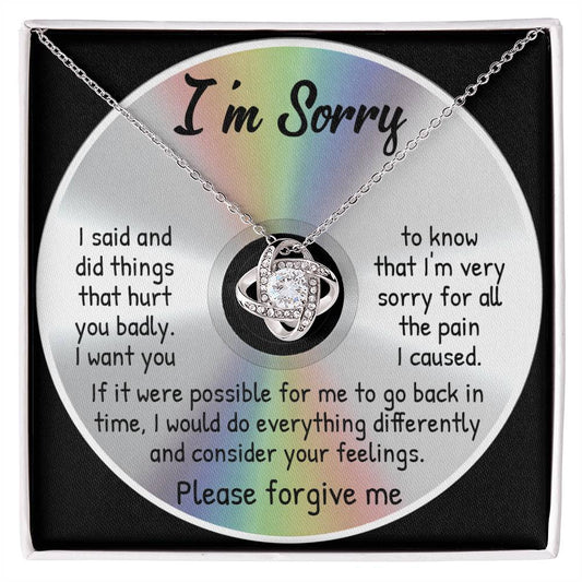 The Sorry-Back In Time - Love Knot Necklace, featuring a crystal pendant, is displayed on a card against a CD image. Crafted from 14k white gold over stainless steel and embellished with cubic zirconia crystals, the card bears an apology message: "I'm Sorry... Please forgive me.