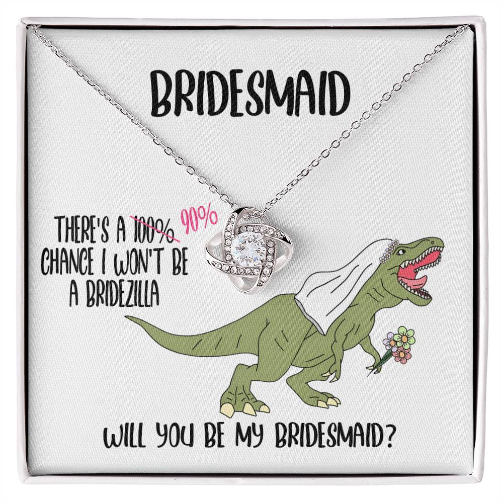 A bridesmaid proposal necklace box featuring a dinosaur with a veil and a stunning To Bridesmaid, Will You Be - Love Knot Necklace in white gold. The text reads, "There's a 90% chance I won't be a bridezilla. Will you be my bridesmaid?