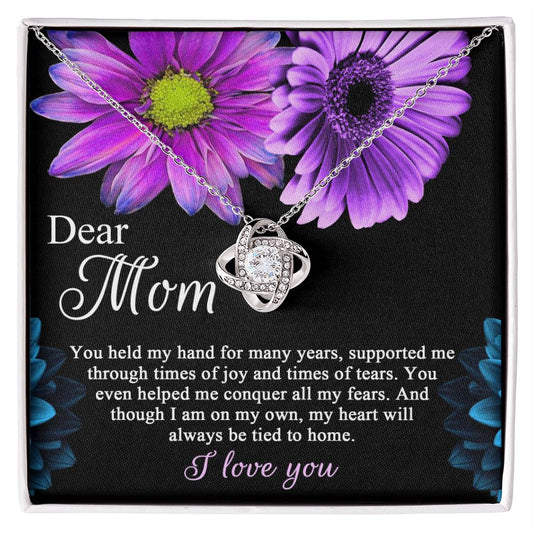 To Mom, Held My Hand - Love Knot Necklace pendant on a chain lying on a black cloth with a printed message to mom and purple flowers in the background.