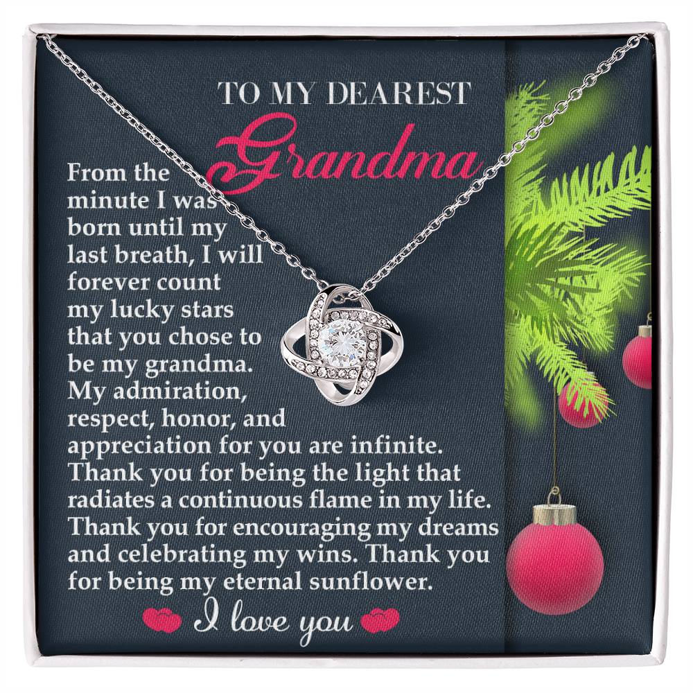 The Grandma-Eternal Sunflower - Love Knot Necklace, featuring a white gold finish and a sparkling cubic zirconia pendant, is elegantly packaged in a box. It includes a heartfelt message to a grandmother, adorned with ornaments and the touching phrase "I love you.