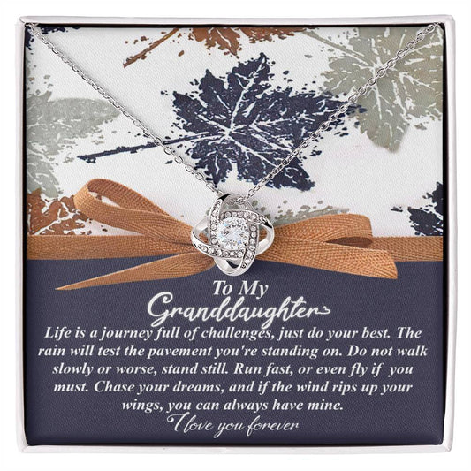 The Granddaughter-Have Mine - Love Knot Necklace, featuring a lustrous gold finish and dazzling cubic zirconia crystals, is beautifully showcased on a floral backdrop. A heartfelt message reads: "To My Granddaughter," accompanied by words of encouragement and love.