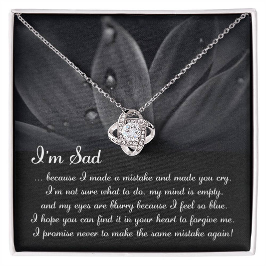 The "Sorry-Feel So Blue - Love Knot Necklace," featuring a stunning knot pendant adorned with cubic zirconia crystals, is displayed over a card with an apologetic message starting with "I'm Sad" and set against a floral background. This personalized gift beautifully combines elegance and heartfelt sentiment.