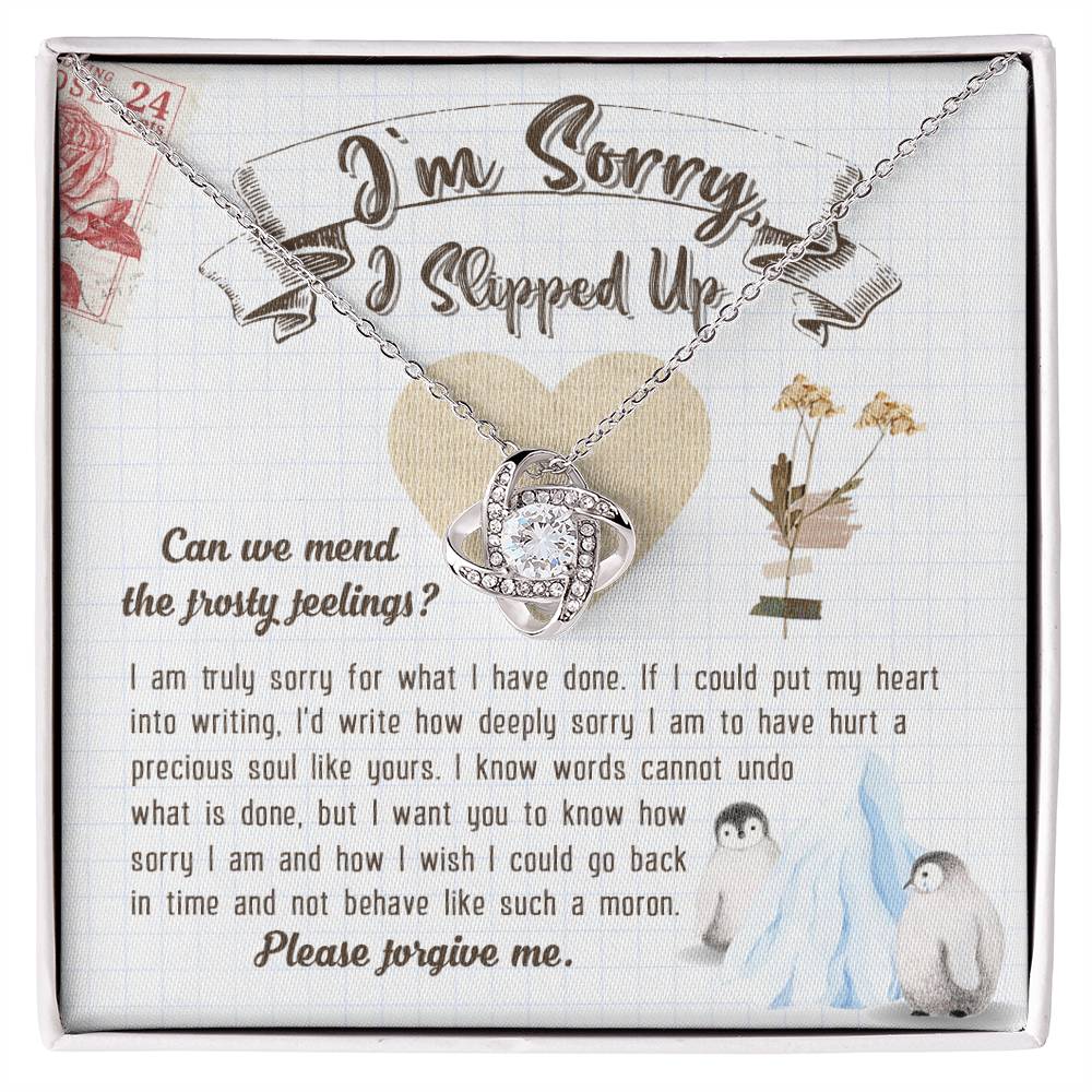 The Sorry-Precious Soul - Love Knot Necklace, featuring an intertwined heart pendant adorned with cubic zirconia crystals, is beautifully displayed in a box. The box comes with an apology note and charming illustrations of flowers, mail stamps, and penguins on a grid-patterned background.