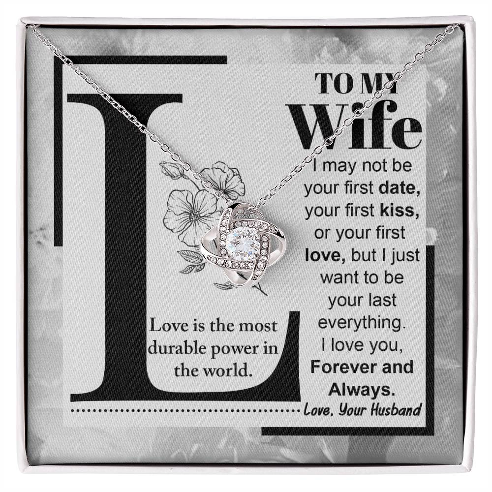 A "to Wife, Love Is - Love Knot Necklace" with a knot design is displayed on a card featuring a printed message to a wife about lasting love, signed from a husband. The piece sparkles with cubic zirconia crystals and boasts an elegant gold finish.