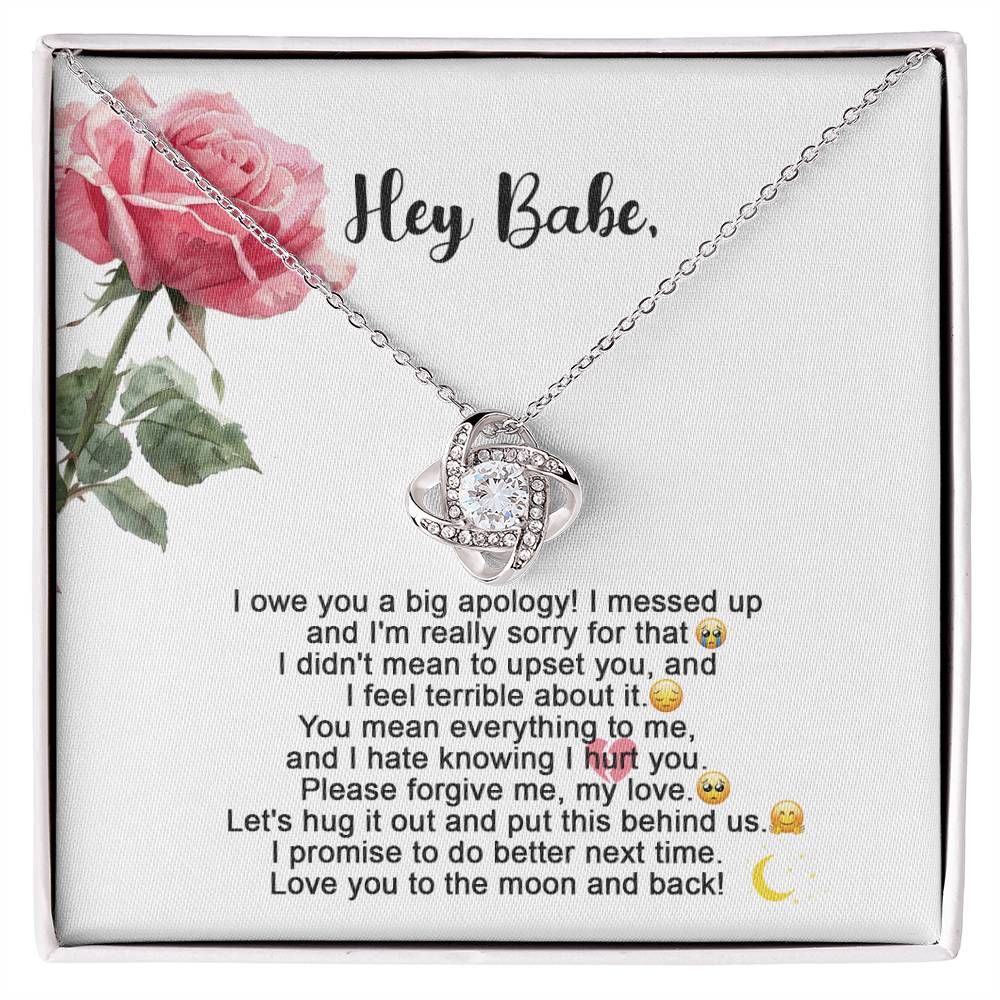 The "Sorry-Hug It Out - Love Knot Necklace" features an 18k yellow gold pendant adorned with small cubic zirconia crystals. It comes on a card decorated with roses, accompanied by the message "Hey Babe" and an apology note.