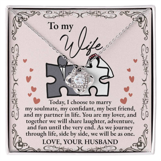 A "To Wife, Be As One - Love Knot Necklace" with intertwined hearts is placed on a card adorned with cubic zirconia crystals, expressing love and commitment from a husband to his wife. The card features a heart and puzzle piece design, available in white gold or yellow gold finish.