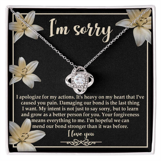 The "Sorry-The Last Thing - Love Knot Necklace" showcases a love knot pendant against a striking black background adorned with white flowers. It's accompanied by an apology message that reads, "I'm sorry. I apologize for my actions... I'm hopeful we can mend our bond stronger," and is beautifully finished with white gold and shimmering cubic zirconia crystals.