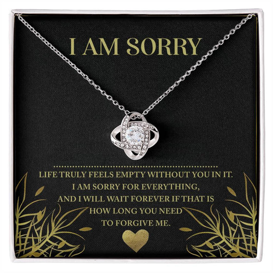 A Sorry-Without You - Love Knot Necklace in silver, featuring an intricate interwoven knot pendant adorned with sparkling cubic zirconia, is showcased against a black background. The packaging bears a heartfelt message in white and gold lettering: "I AM SORRY" at the top, followed by, "Life truly feels empty without you in it. I am sorry for everything, and I will wait forever if that is how long you need.