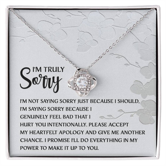 Introducing the Sorry-Feel Bad - Love Knot Necklace, featuring a pendant on a card with an apology message: "I'm truly sorry. I'm not saying sorry just because I should..." and continues with more heartfelt text. The necklace showcases a sparkling cubic zirconia crystal set in your choice of white gold or yellow gold finish, all against a light floral design background.