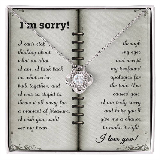 The "Sorry-What An Idiot - Love Knot Necklace" made of 14k white gold is displayed on an open card. The pendant, adorned with cubic zirconia crystals, hangs gracefully, and the card holds a heartfelt apology message.