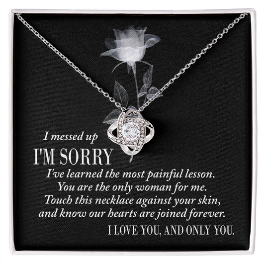 A silver Sorry-Painful Lesson - Love Knot Necklace on a dark background, adorned with shimmering cubic zirconia crystals. The text includes an apology, a statement of love, and an affirmation of unity.