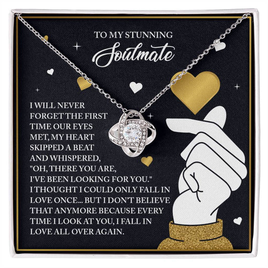 The Soulmate-Look At You Love Knot Necklace, a silver piece with a gold finish, rests on a black and gold backdrop. It features romantic motifs of hearts and an arrow, and its cubic zirconia pendant symbolizes endless love.