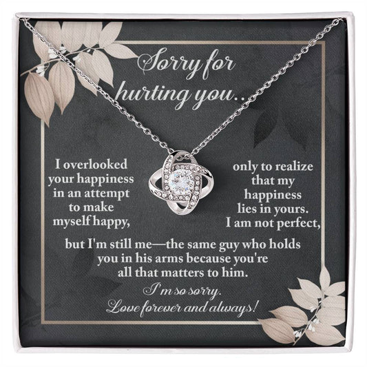 The Sorry-Lies In Yours - Love Knot Necklace, a stunning piece embellished with cubic zirconia crystals, is showcased against a dark-themed backdrop adorned with floral designs. The background beautifully conveys an apology message infused with love and regret.