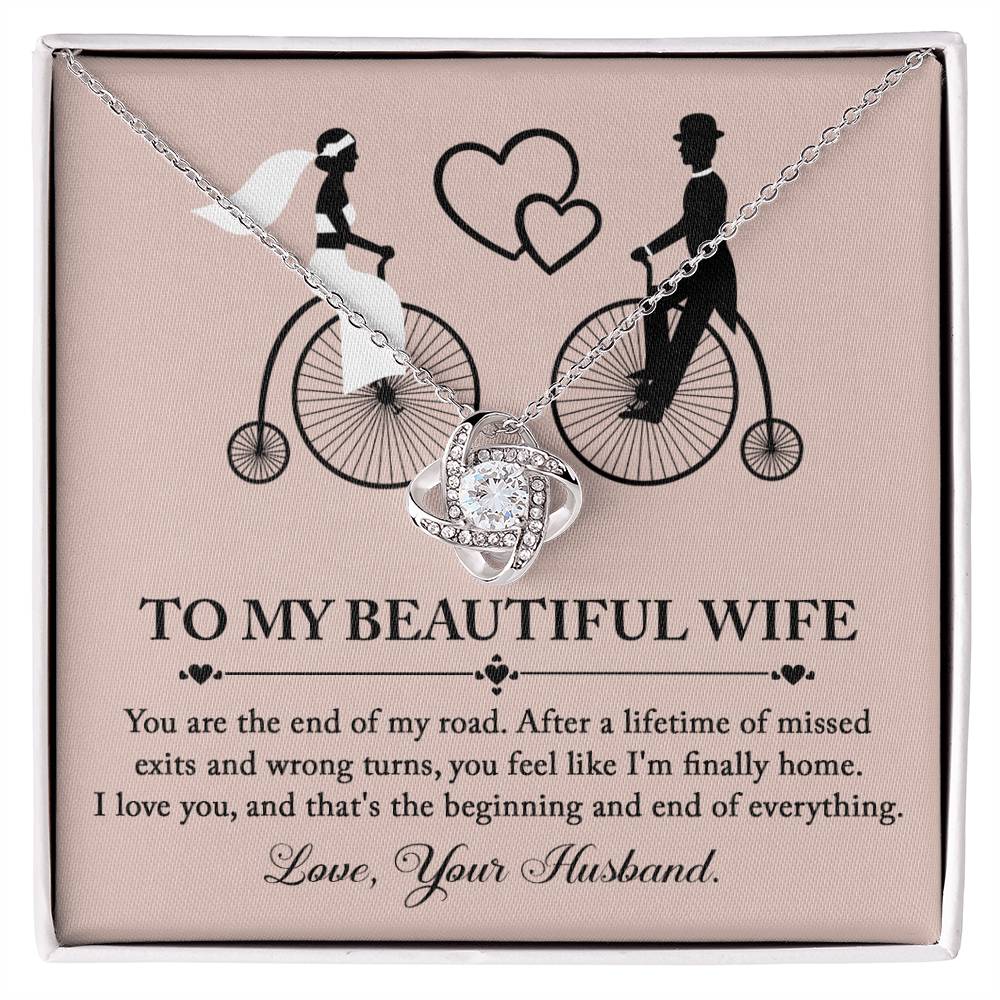 To Wife, I'm Finally Home - Love Knot Necklace with intertwined circle pendant on a card featuring an illustration of a couple on bicycles. The necklace, adorned with cubic zirconia crystals and a stunning gold finish, comes with a card that reads: 'To my beautiful wife' followed by a romantic message and ending with 'Love, Your Husband.'