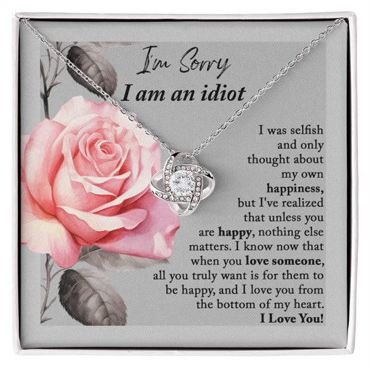 The "Sorry-Love Someone - Love Knot Necklace" is an elegant silver piece with a heart-shaped pendant, adorned with sparkling cubic zirconia crystals. It rests on a beautifully designed box featuring a pink rose and a heartfelt apology note, making it truly a classic gift.