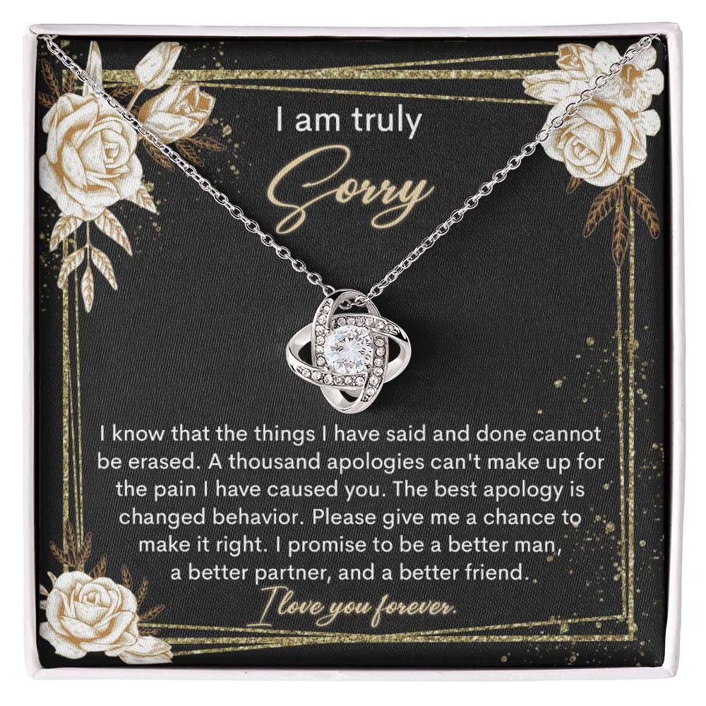 The Sorry-Cannot Be Erased - Love Knot Necklace, adorned with shimmering cubic zirconia crystals and available in a choice of white gold or yellow gold finish, is displayed against a backdrop of a black card bearing an apology message that reads, "I am truly sorry" and "I promise to be a better man, a better partner, and a better friend.