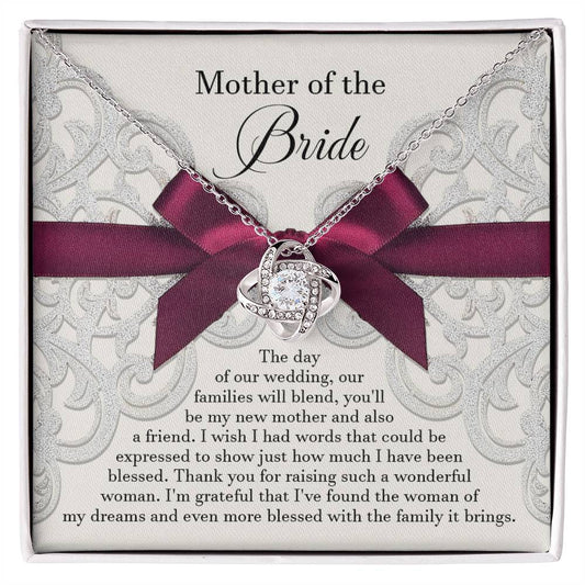 A To Bride's Mom, My New Mother - Love Knot Necklace with a heart pendant, crafted from 14k white gold and adorned with cubic zirconia crystals, is displayed on a decorative card with a maroon ribbon. The card reads, "Mother of the Bride" and contains a heartfelt message of gratitude.