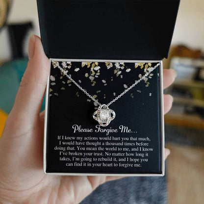 A hand holding an open black box reveals a stunning Sorry-Broken Your Trust - Love Knot Necklace adorned with shimmering cubic zirconia crystals. The intertwined design elegantly cradles a heartfelt message that reads, "Please Forgive Me," conveying deep regret and remorse for past actions.