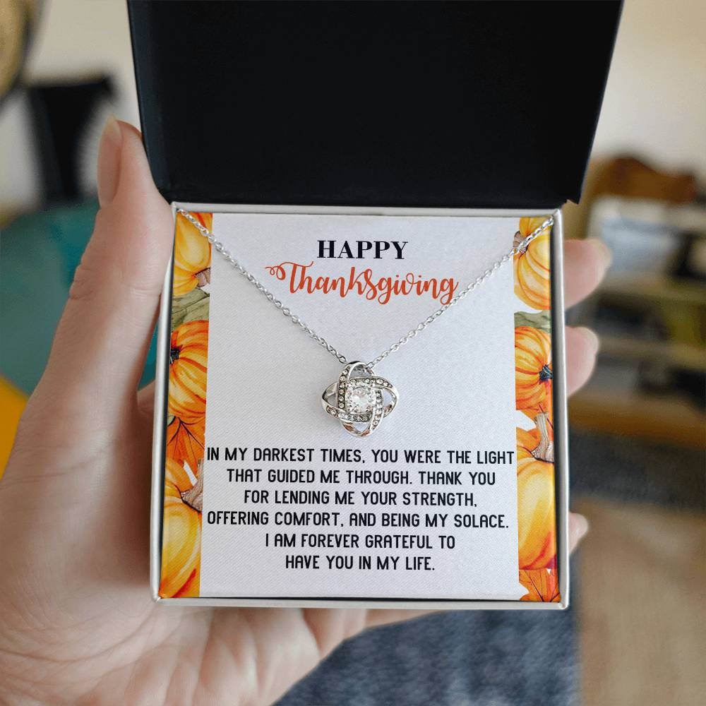 A hand holds an open jewelry box containing the Thanksgiving-Guided Me Through - Love Knot Necklace, adorned with sparkling cubic zirconia crystals and accompanied by a card expressing gratitude and appreciation.