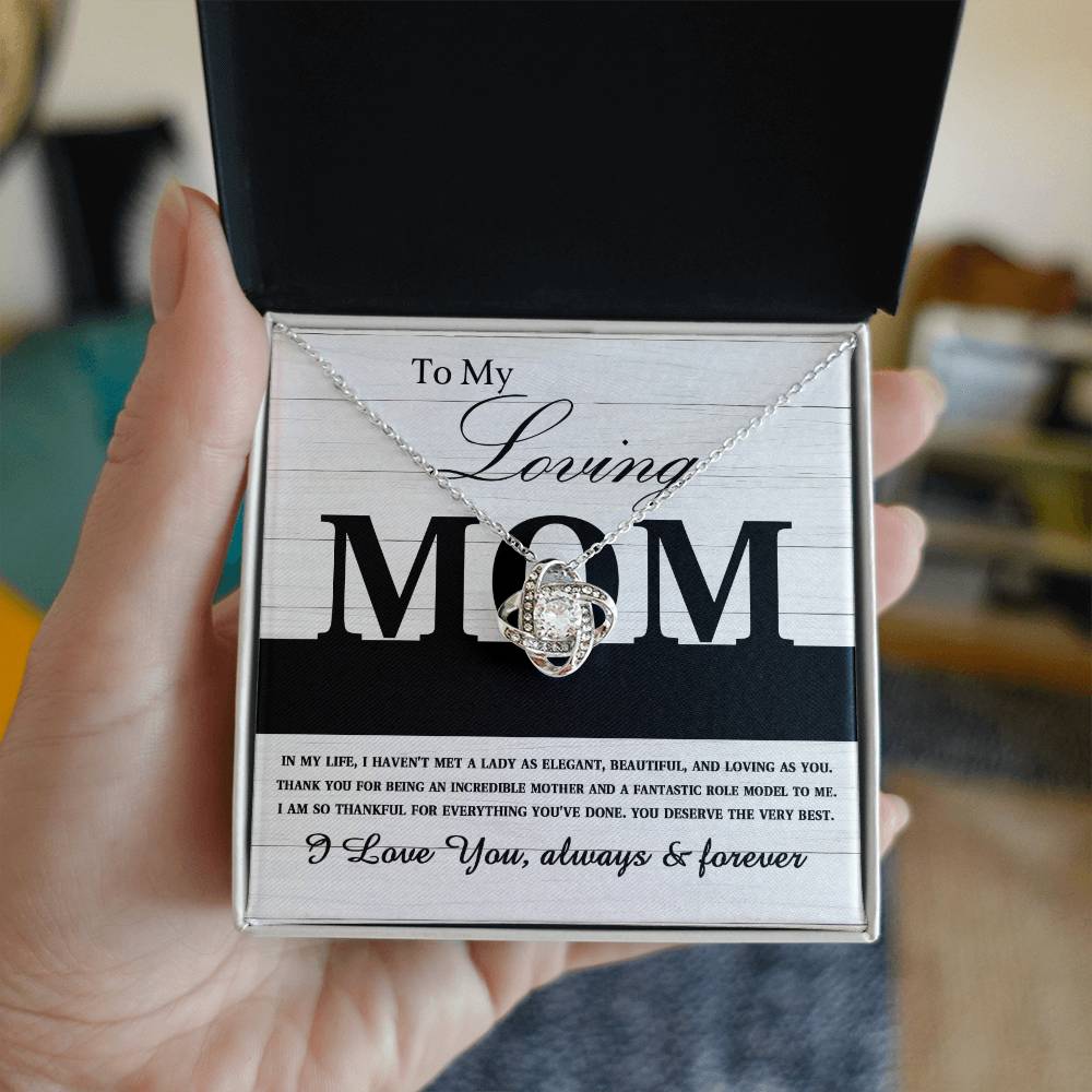 A hand holds an open jewelry box displaying a "To Mom, Loving As You - Love Knot Necklace" adorned with cubic zirconia crystals and a message that reads, "To My Loving Mom. I love you, always & forever.