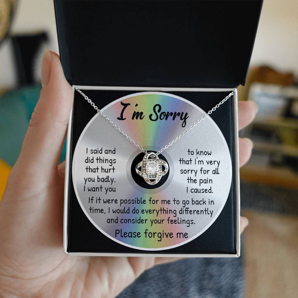 A person holding a box containing a "Sorry-Back In Time - Love Knot Necklace" adorned with cubic zirconia crystals and accompanied by a card that reads, "I'm Sorry," along with a heartfelt apology message for the past hurt caused.