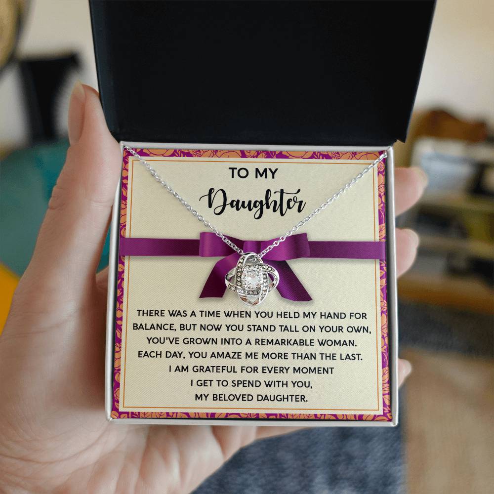 A hand holds an open gift box containing a Daughter-Spend With You - Love Knot Necklace, adorned with cubic zirconia crystals. The card inside has a heartfelt message addressed "To My Daughter," making it the perfect token of love and appreciation.