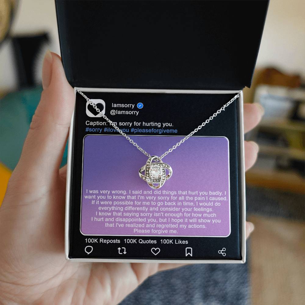 A stunning "Sorry-Hurt You Badly - Love Knot Necklace" in 14k white gold, adorned with cubic zirconia crystals, is displayed in front of a printed Instagram post that includes an apology message and hashtags like #sorry and #iloveyou.