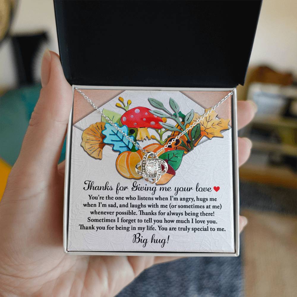 A hand holds an open box revealing the exquisite Thanksgiving-Big Hug - Love Knot Necklace, featuring a gleaming gold finish. The necklace rests on a card embellished with vibrant leaf and mushroom illustrations, accompanied by a heartfelt message of gratitude and love, making this gift truly special.