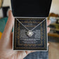 A hand holds an open gift box containing the "To Mother-In-Law, Through My Eyes - Love Knot Necklace" adorned with premium cubic zirconia. The box features a heartfelt message expressing gratitude to a mother-in-law, making this personalized gift truly special.
