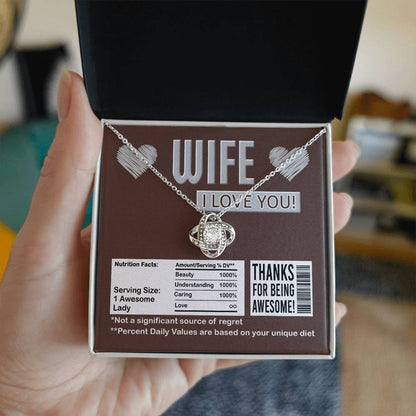 A To Wife, Nutrition Facts - Love Knot Necklace in a gift box that reads, "WIFE I LOVE YOU!" and "Thanks for being awesome!" It humorously lists nutritional facts like "Beauty 1000%" and "Not a significant source of regret," featuring sparkling cubic zirconia crystals with a stunning gold finish.