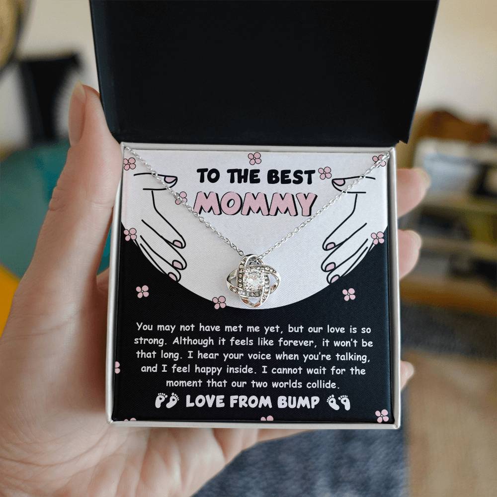 A hand holds an open box revealing the "To Mom To Be, Two Worlds Collide - Love Knot Necklace" adorned with cubic zirconia crystals, along with a card that reads "To the best Mommy" and a heartfelt message from an unborn baby, symbolizing their unbreakable bond.