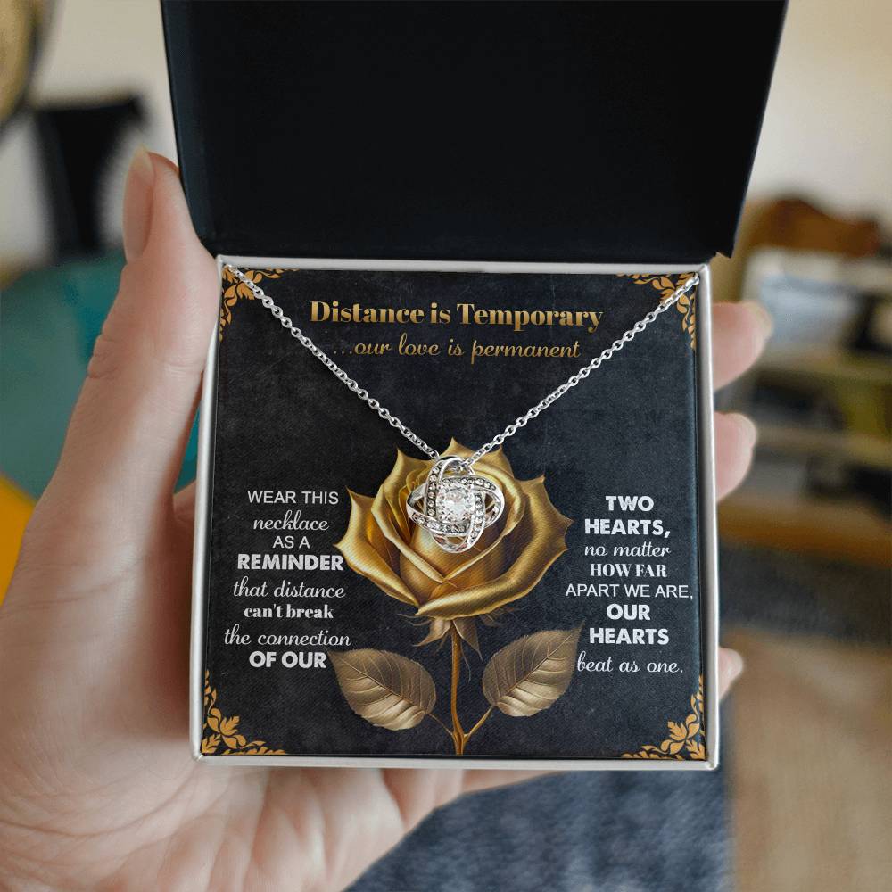 A hand opens a jewelry box to reveal the Soulmate-Love Is Permanent Love Knot Necklace adorned with cubic zirconia crystals. The box showcases a gold rose design and the phrase "Distance is Temporary, our love is permanent," making it an ideal personalized gift with heartfelt inscriptions.