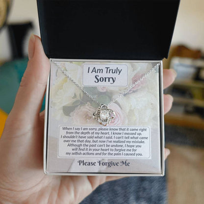 A person holds an open box containing a Sorry-What I Said - Love Knot Necklace with a silver pendant alongside a printed apology note. The text reads "I Am Truly Sorry" and includes a heartfelt apology message ending with "Please Forgive Me.