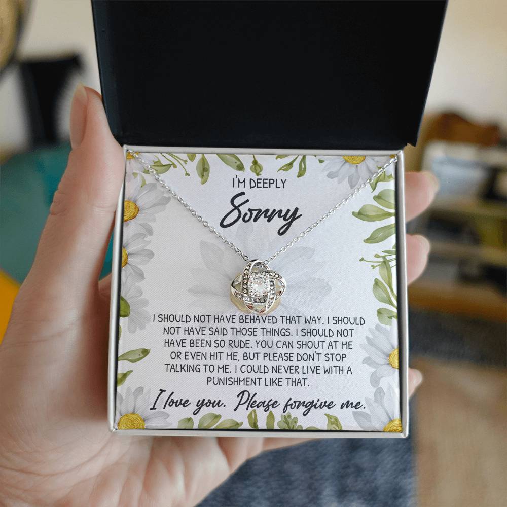 A hand holds an open box revealing the Sorry-Please Don’t Top - Love Knot Necklace, featuring a heart-shaped pendant adorned with cubic zirconia crystals. The box's interior showcases a floral background and includes a printed apology message expressing deep regret and asking for forgiveness.