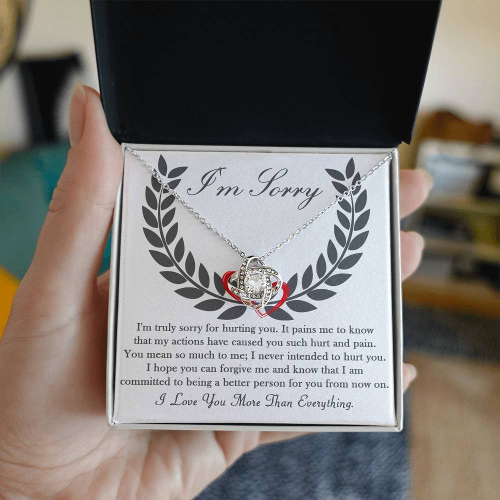 Close-up of a hand holding an open box containing the "Sorry-From Now On - Love Knot Necklace," which features a silver necklace with a cubic zirconia crystal-studded ring pendant. The box includes an apology message and the text "I'm Sorry" at the top, set against a blurred background.