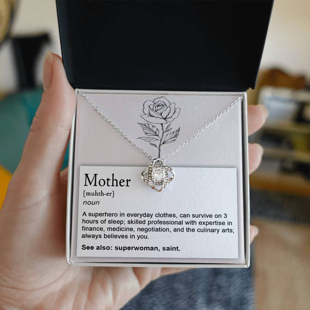 A hand holds an open jewelry box containing the "To Mom, Superhero - Love Knot Necklace" adorned with cubic zirconia crystals and a card defining "Mother," highlighting attributes such as surviving on little sleep, mastering various professions, and always believing in you.