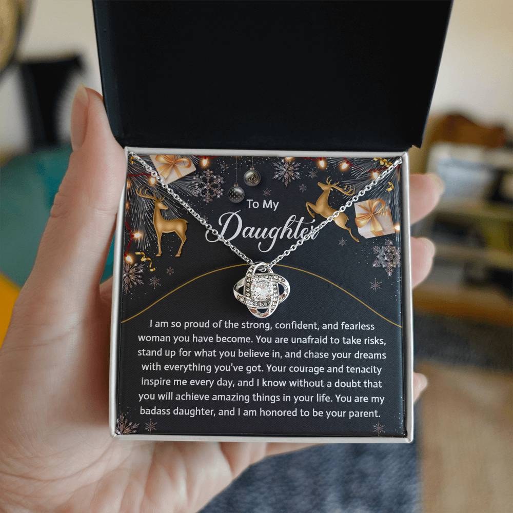 A hand holds an open gift box revealing the Daughter-Amazing Things - Love Knot Necklace, embellished with cubic zirconia crystals. The card inside delivers a personalized message of pride and encouragement to a daughter.