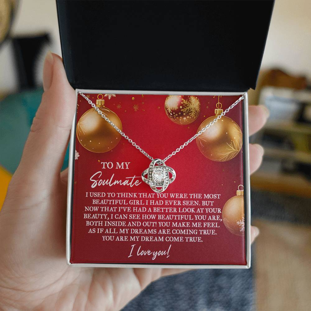 A hand reveals an open jewelry box containing the Soulmate-Your Beauty - Love Knot Necklace with a round pendant inside. This necklace, embellished with Cubic Zirconia, shines beautifully against its white gold finish. The lid of the box features a heartfelt holiday-themed message dedicated to a soulmate.