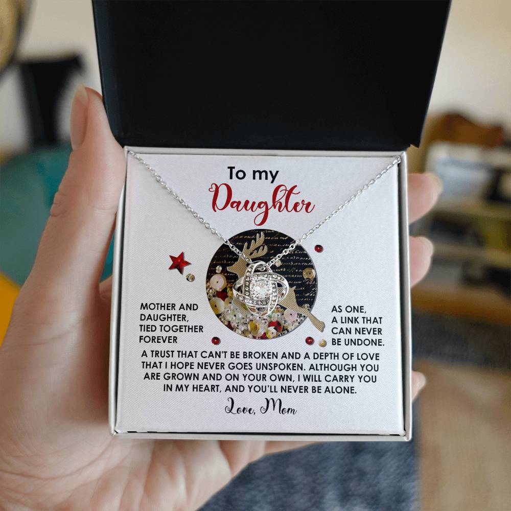 A hand holds an open gift box containing the Daughter-Forever As One - Love Knot Necklace, embellished with cubic zirconia crystals. The box lid showcases a heartfelt message from a mother to her daughter, making it a truly unique gift.