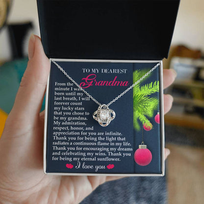 A hand holds an open gift box revealing a stunning Grandma-Eternal Sunflower - Love Knot Necklace with a white gold finish. The inside lid features an appreciation message addressed to "Grandma," adorned with holiday decorations that perfectly complement the elegant cubic zirconia centerpiece.