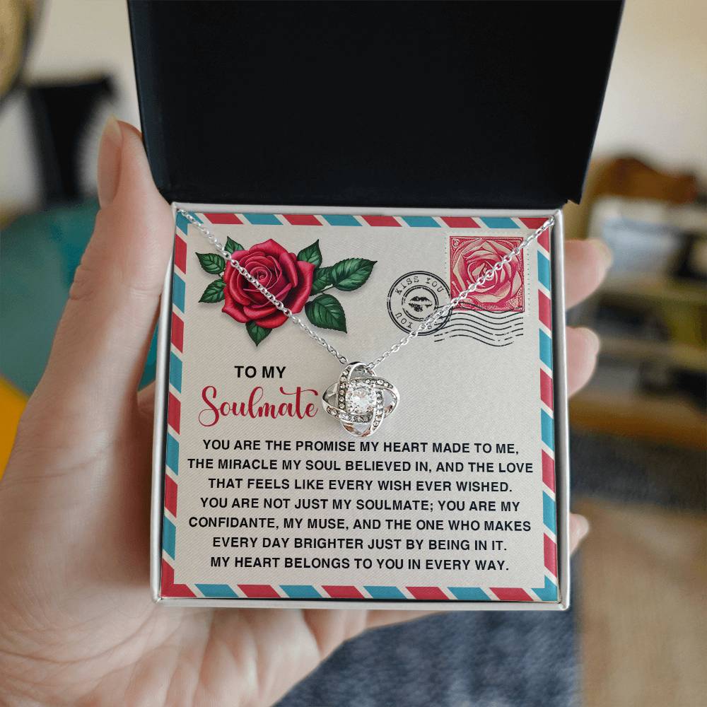 A person opens a box revealing the stunning "Soulmate-Made To Me - Love Knot Necklace" adorned with cubic zirconia crystals, alongside a card elegantly featuring a message to their soulmate, highlighted by a rose and decorative text.