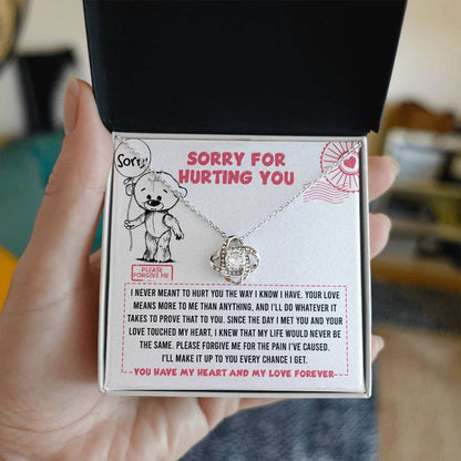 A hand holds an open jewelry box with the Sorry-Every Chance - Love Knot Necklace inside. The 14k white gold necklace is adorned with shimmering cubic zirconia crystals. The box has a printed apology message and an illustration of a sad teddy bear saying, "Sorry for hurting you.