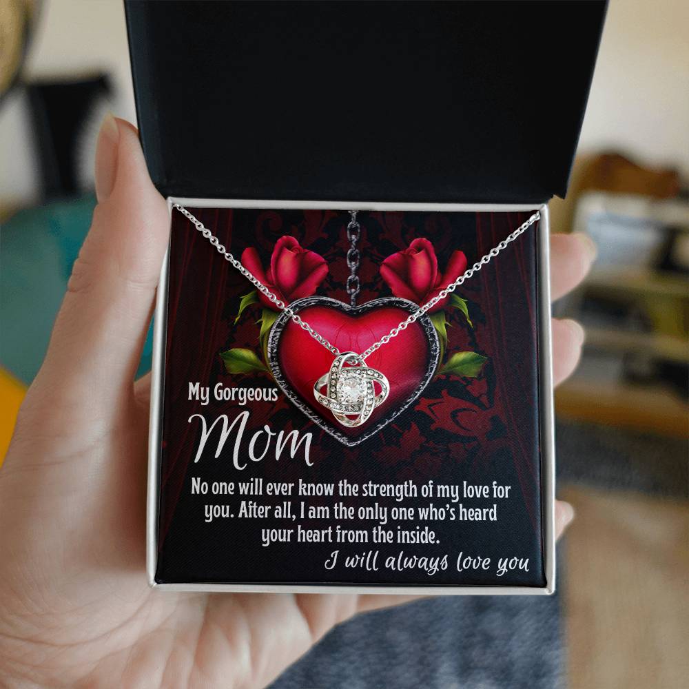 A hand holds an open jewelry box containing the [Almost Sold Out] To Mom, Heard Your Heart - Love Knot Necklace, adorned with sparkling cubic zirconia crystals. A card inside reads: "My Gorgeous Mom, No one will ever know the strength of my love for you. After all, I am the only one who's heard your heart from the inside.