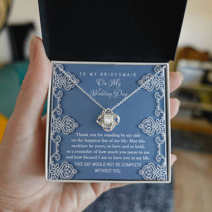 To Bridesmaid, By My Side - Love Knot Necklace with interlocking heart design, adorned with sparkling cubic zirconia crystals, displayed in a box with a heartfelt message for the bridesmaid. The message expresses gratitude for her unwavering support on the wedding day.