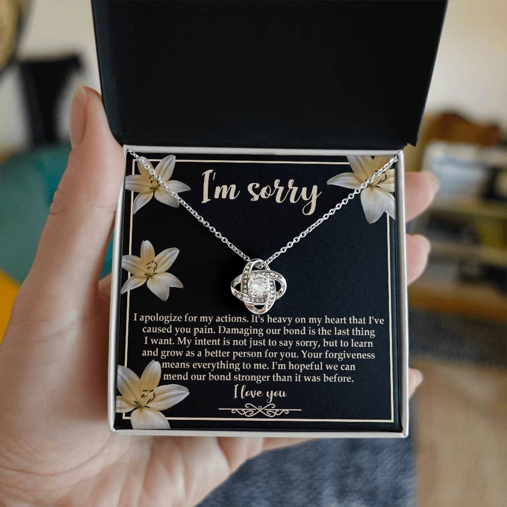 The "Sorry-The Last Thing - Love Knot Necklace" showcases a love knot pendant against a striking black background adorned with white flowers. It's accompanied by an apology message that reads, "I'm sorry. I apologize for my actions... I'm hopeful we can mend our bond stronger," and is beautifully finished with white gold and shimmering cubic zirconia crystals.