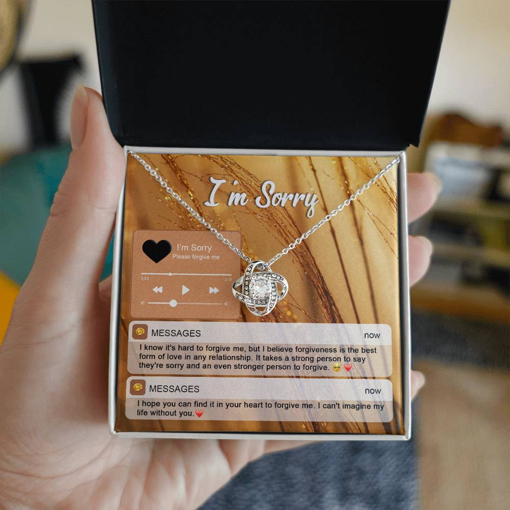 The "Sorry, Person To Forgive - Love Knot Necklace" features a gold finish and sparkling cubic zirconia crystals. It is presented on a card adorned with musical notes and icons that reads "I'm Sorry," along with heartfelt messages of apology and pleas for forgiveness.