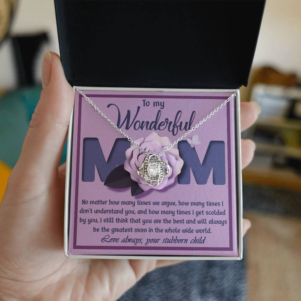 A hand holds an open jewelry box with the "To Mom, The Greatest Mom - Love Knot Necklace" adorned with cubic zirconia crystals. The box lid features a heartfelt message to a mom, acknowledging misunderstandings, scolding, and expressing love from a child—a perfect personalized gift.