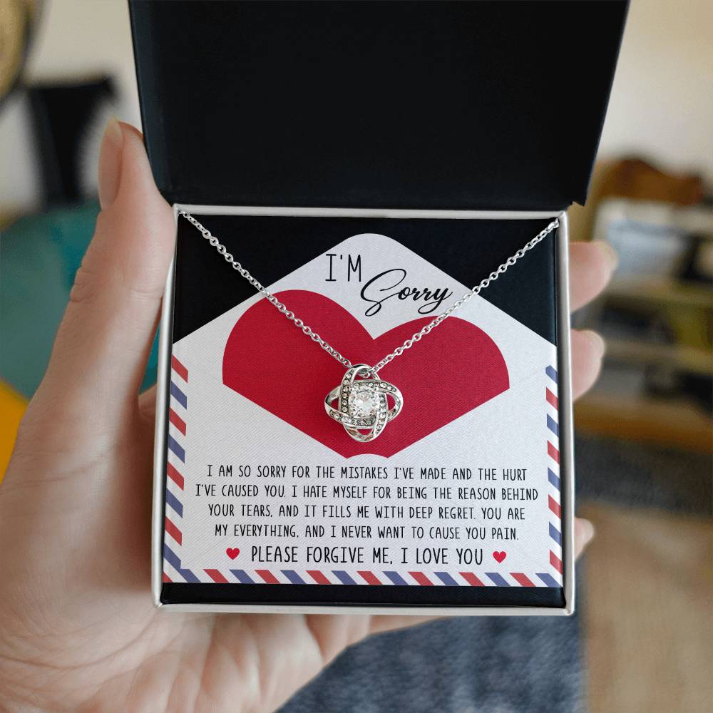 A hand holds an open gift box containing a stunning Sorry-Behind Your Tears - Love Knot Necklace adorned with cubic zirconia crystals, accompanied by an apology note that reads 'I'm sorry' and a heartfelt message expressing regret and asking for forgiveness.